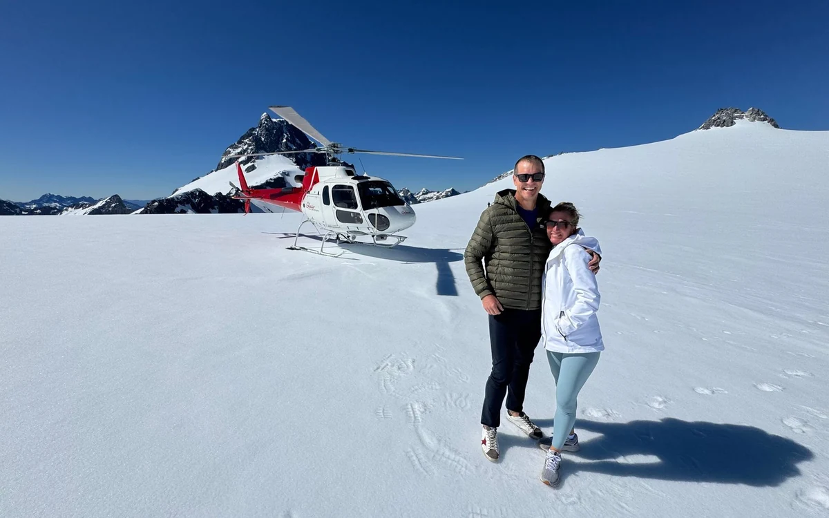 Passengers standing on a snow-covered peak beside a helicopter with mountain ranges behind them