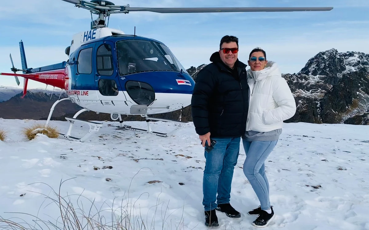 Two passengers standing on a mountain ridge beside a helicopter with the valley far below