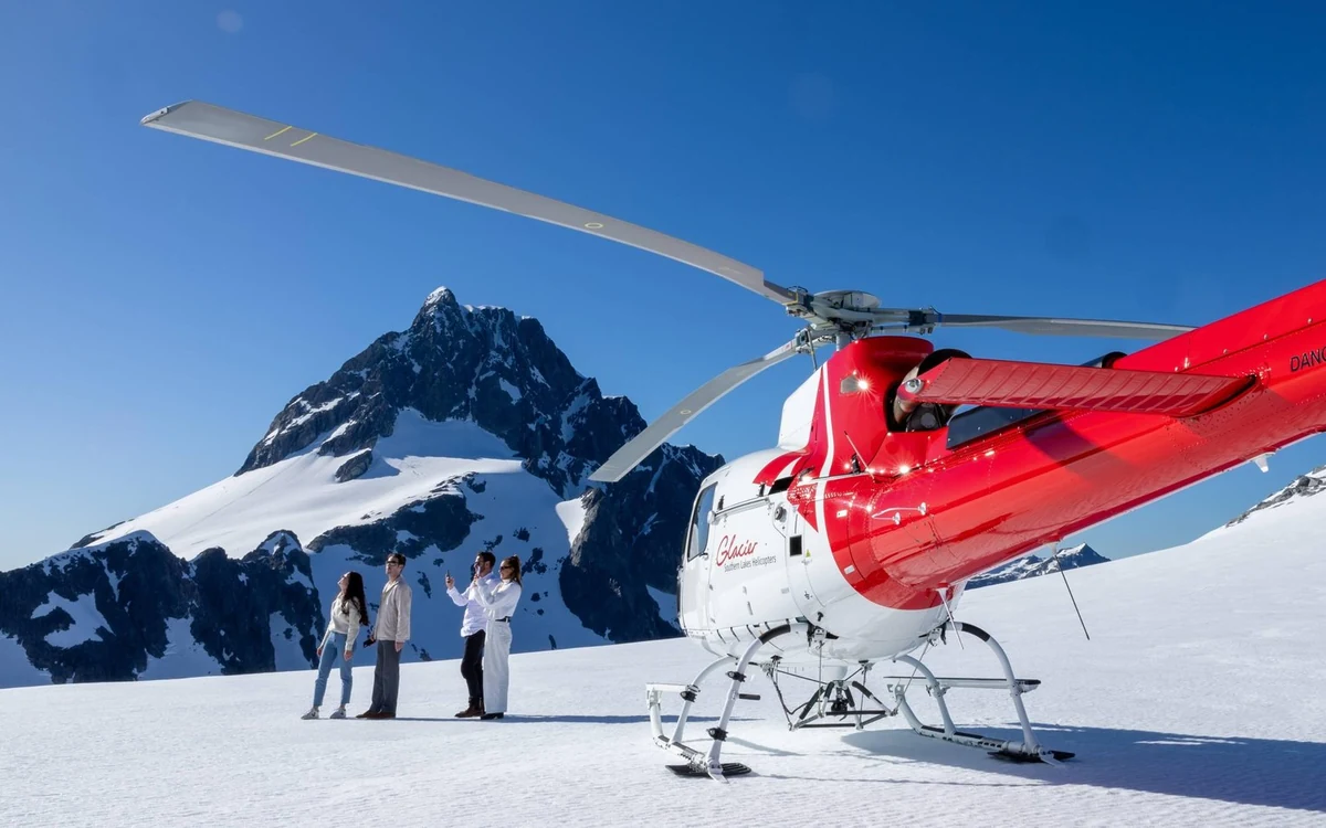A red helicopter flying along a snow-covered ridge in the Southern Alps