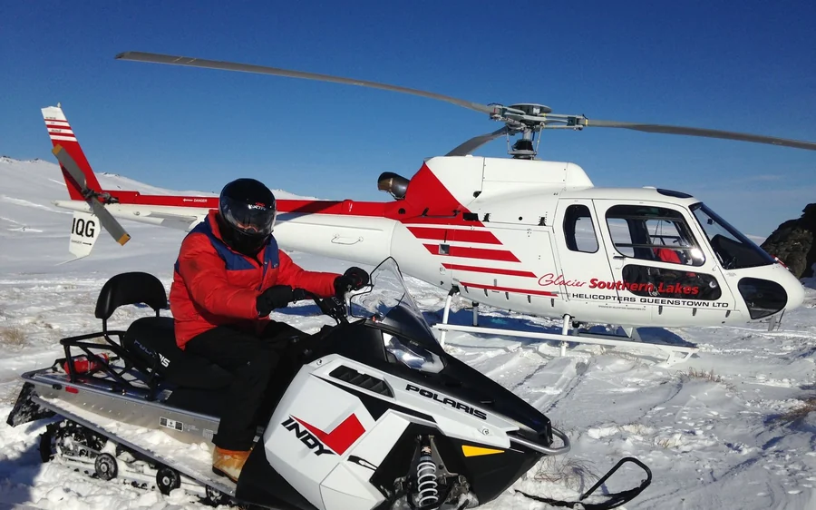 Snowmobiles parked on a high snow plateau with a helicopter nearby